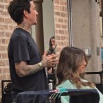 A male hair educator with tattoos on his neck and arms gestures while explaining a technique to a student during the DOM DOM advanced education class.