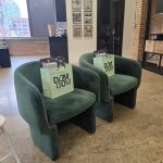 Two plush forest green velvet chairs in a modern salon loft, each holding a lime green "DOM DOM" gift bag.